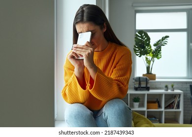 Depressed Woman Hiding Face Behind Smart Phone While Sitting On The Window Sill At Home