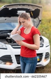 Depressed Woman Looking At Her Broken Car And Reading Car Owner Manual