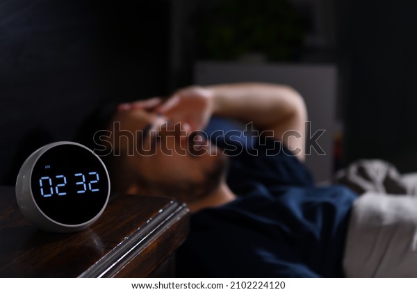 Depressed young Asian man lying in bed cannot sleep\
from insomnia. focus on\
clock