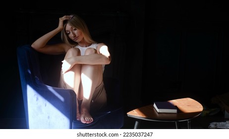 A depressed young beautiful woman sits alone in an armchair in the dark, hugs her knees and is sad. Loneliness sad woman thinks about grief, problems of life. Negative emotions and mental health - Shutterstock ID 2235037247