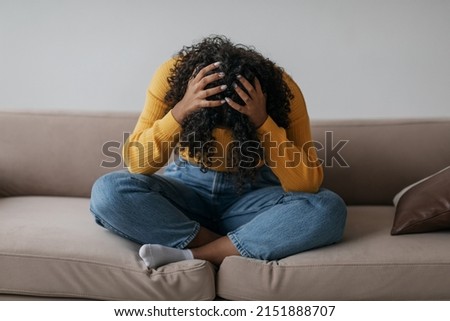 Depressed young black woman sitting cross legged on sofa with head in her hands at home. African American female patient feeling stressed, suffering from PTSD or having psychological problem