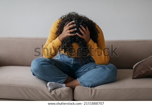 Depressed young black woman sitting cross legged\
on sofa with head in her hands at home. African American female\
patient feeling stressed, suffering from PTSD or having\
psychological problem