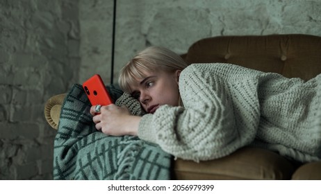 Depressed Young Blonde Woman Scrolling Through Her Phone, Refusing To Get Up From The Sofa. Beautiful Girl Looking Sad While Laying On The Couch On Her Smartphone.