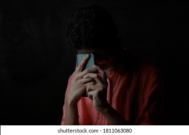 A Depressed Young Boy Placing His Mobile Phone On His Forehead. One Light Studio Shot With Shadow