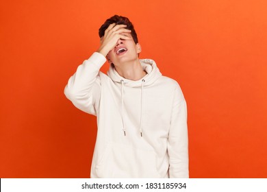 Depressed young man in casual style white hoodie closing eyes with arm, making facepalm gesture, loser, forget about something important. Indoor studio shot isolated on orange background
