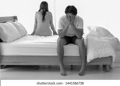 Depressed Young Man Sitting On The Edge Of The Bed And Holding The Head With Hand,having Relationship Problem With His Girlfriend.Woman Sitting And Turned Her Back On Him In Bedroom. Black And White.