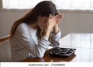 Depressed young mixed race woman sitting at table with chocolate sweets, feeling desperate alone at home. Addicted to candies stressed millennial girl suffering from eating disorder, anorexia concept.