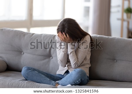 Depressed young woman covering face by hands, crying alone at home, upset girl sitting on sofa, feeling unhappy after quarrel or breakup, despair and lonely, psychological problem concept