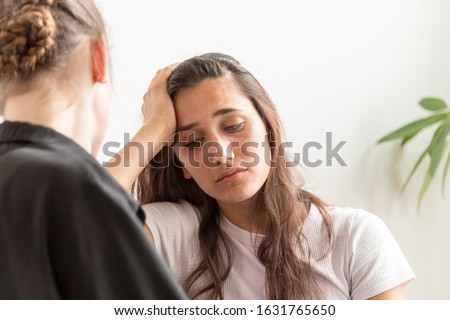 Depressed young woman with hand on forehead with girlfriend sitting near. Medium close up.