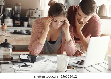 Depressed young woman having bad headache holding her temples feeling stressed by lack of money to pay out family debts, her puzzled husband standing next to her, trying to come up with solution