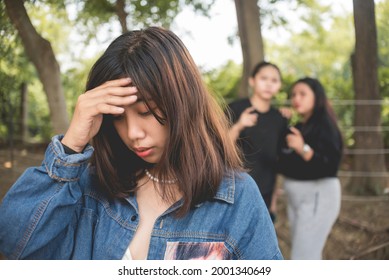 A depressed young woman holds her forehead in shame while 2 women talk and gossip behind her back. Teenage bullying or backstabbing.