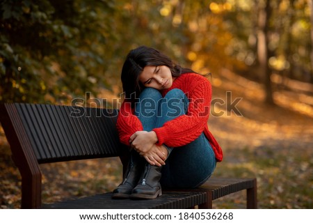 Depressed young woman hugging her knees while sitting on bench at yellow autumn park. Millennial lady feeling stressed or unhappy, suffering from loneliness or seasonal affective disorder, outdoors