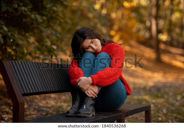 Depressed young woman hugging her knees while\
sitting on bench at yellow autumn park. Millennial lady feeling\
stressed or unhappy, suffering from loneliness or seasonal\
affective disorder,\
outdoors