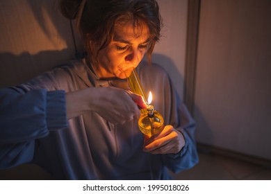 Depressed Young Woman Sitting On The Floor At Dark, Smoking Pot Using Bong; Woman Vaping Marijuana With Bong