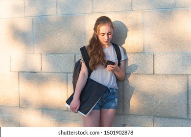 Depressed/Sad Teen Girl Leaning Against High School Wall During Sunset While Wearing A Backpack And Holding Binders/smartphone.