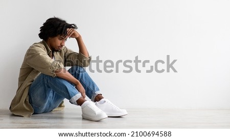 Depression Concept. Portrait Of Upset Black Guy Sitting On Floor Near White Wall Indoors, Anxious Young African American Man Touching Forehead And Thinking About Something, Panorama With Free Space