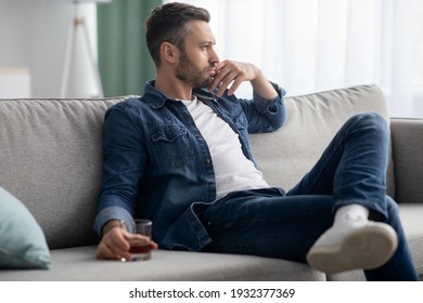 Depression, crisis concept. Sad middle-aged bearded man sitting on sofa, drinking alcohol at home, feeling lonely and sick, holding glass of whiskey and looking through the window, copy space