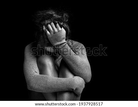 Depression or domestic violence concept: Black and white photo of Sad young woman crying while sitting in dark room with an attitude of sadness and boredom with her legs together and hugged.