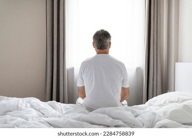 Depression, midlife crisis, mental breakdown. Back view of grey-haired middle aged man wearing pajamas sitting alone on bed after waking up at home, looking through window, copy space - Powered by Shutterstock