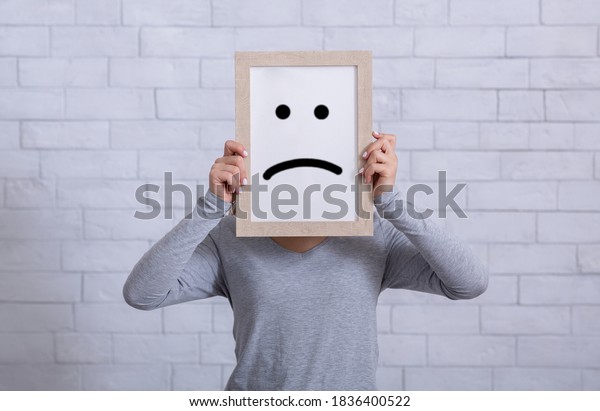 Depression, sadness, negative emotions\
concept. Young woman holding picture frame with sad emoticon in\
front of her face. Millennial lady expressing pessimism,\
loneliness, grief or\
frustration