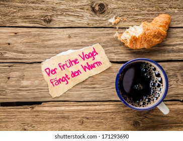 Der fruehe Vogel faengt den Wurm - the early bird catches the worm - an inspirational message on a torn piece of paper alonside an early breakfast of espresso coffee and half eaten croissant