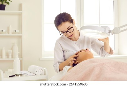 Dermatologist, skin therapist, or beautician looking at patient's face under magnifying lamp. Cosmetologist examining woman's skin before doing facial, removing blackheads, or cleaning clogged pores - Shutterstock ID 2141559685