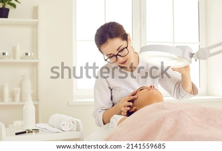 Dermatologist, skin therapist, or beautician looking at patient's face under magnifying lamp. Cosmetologist examining woman's skin before doing facial, removing blackheads, or cleaning clogged pores