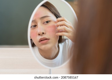 Dermatology, puberty asian young woman, girl looking into mirror, allergy presenting an allergic reaction from cosmetic, red spot or  rash on face. Beauty care from skin problem by medical treatment. - Shutterstock ID 2087585167
