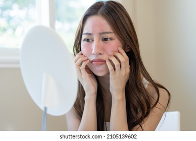 Dermatology, puberty asian young woman, girl looking into mirror, allergy presenting an allergic reaction from cosmetic, red spot or  rash on face. Beauty care from skin problem by medical treatment. - Shutterstock ID 2105139602