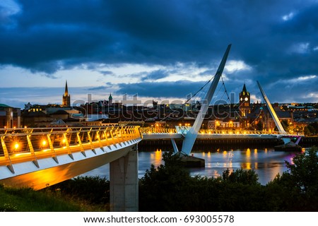 Derry, Ireland. Illuminated Peace bridge in Derry Londonderry in Northern Ireland with city center at the background. Night cloudy sky, reflection in the river.