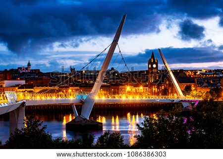 Derry, Northern Ireland. Illuminated Peace bridge in Derry Londonderry in Northern Ireland with city center at the background. Night cloudy sky, reflection in the river.