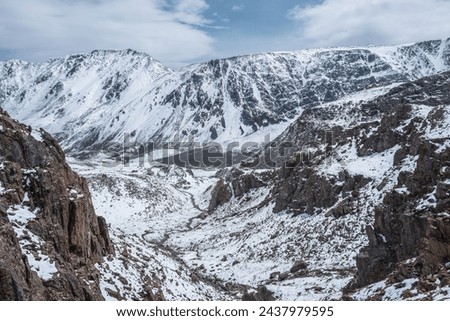 Descent along thin rocky gorge down into snow-covered alpine valley. Landscape from above to snowy stony couloir. White snow on stones in high mountains. Top view to snow-white steep slope among rocks