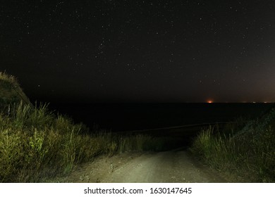 Descent Of A Dirt Road From A Hill To The Night Sea. Sea Coast At Night. Starry Nightscape Over The Sea.