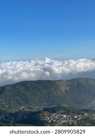 Description: A breathtaking panoramic view of a lush green mountain slope towering above a dense, fluffy white sea of clouds. The landscape features rolling forested hills with patches of exposed earth, leading down to a charming rural village with traditional houses nestled in the valley at the base. The scene is set under a clear, vibrant blue sky, creating a serene and awe-inspiring high-altitude natural landscape, perfect for travel, nature, and environmental concepts.