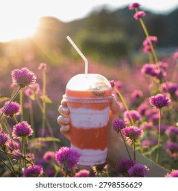 Description: A close-up side view of a woman's hand in a cozy beige sweater holding a clear plastic bottle filled with fresh strawberry yogurt milk, with a beautiful soft-focus pink globe amaranth flower field background.