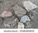 Description: A collection of various stones and concrete fragments scattered on a rough, gravelly surface. The pieces differ in texture, size, and color, ranging from reddish-brown to gray and speckle