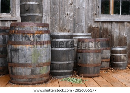 Description should be general, without details, no more than one sentence. A collection of weathered wooden barrels rests on a wooden deck in front of a rustic barn exterior.