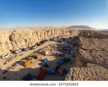 A desert camping site with several cars and tents set up near rocky cliffs under a clear blue sky. People are gathered around, enjoying the outdoor environment and activities. - Powered by Shutterstock