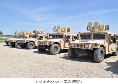 Desert tan US Military armored  High Mobility Multi-Wheeled Vehicles (HMMWV) often used in the wars in Afghanistan and Iraq parked in a gravel lot with a blue sky and green grass in the background. - Shutterstock ID 2138380245