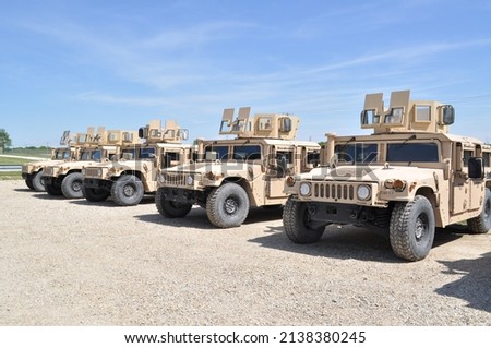 Desert tan US Military armored  High Mobility Multi-Wheeled Vehicles (HMMWV) often used in the wars in Afghanistan and Iraq parked in a gravel lot with a blue sky and green grass in the background.