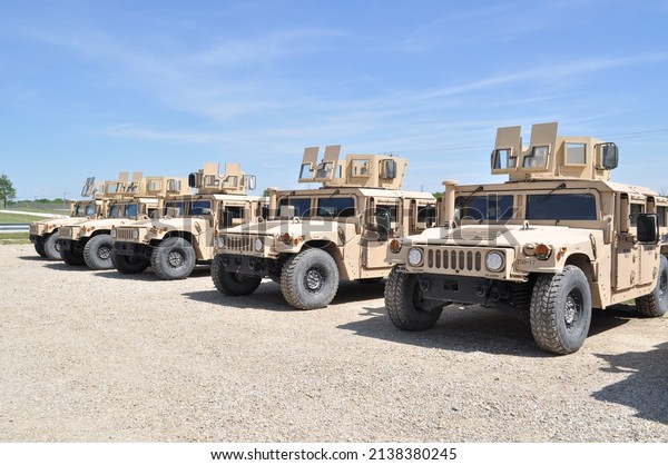 Desert tan US Military armored  High Mobility\
Multi-Wheeled Vehicles (HMMWV) often used in the wars in\
Afghanistan and Iraq parked in a gravel lot with a blue sky and\
green grass in the\
background.