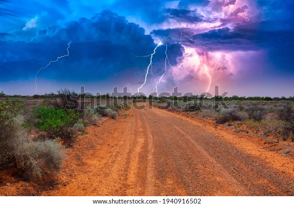 Deserted Australian outback\
landscape with red dirt road towards horizon with bushes in\
roadsides and heavy thunderstorm with white purple lightnings on\
the horizon