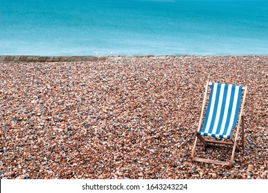 A deserted pebble beach with one empty striped deckchair in lower right foreground. Two seagulls sit on a stone groyne in the background with the sea beyond providing copy space..