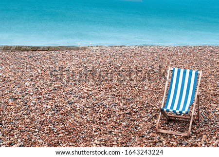 A deserted pebble beach with one empty striped deckchair in lower right foreground. Two seagulls sit on a stone groyne in the background with the sea beyond providing copy space..