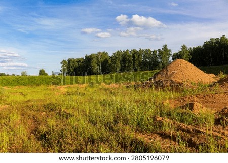 Deserted sand pit featuring massive mounds of earth, scattered gravel, overgrown wild grasses and blooming wildflowers under a bright blue sky with light clouds, scenic reclamation area
