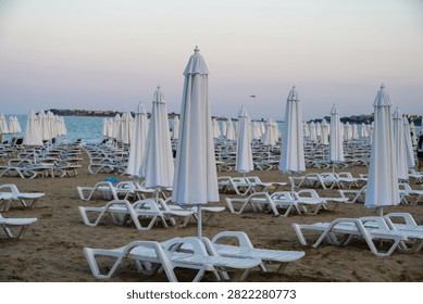 A deserted sandy beach features rows of closed white beach umbrellas and empty lounge chairs under an evening sky.
