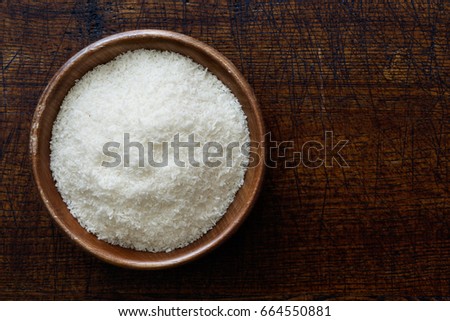 Desiccated coconut in dark wooden bowl isolated on dark brown wood from above.
 