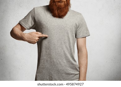 Design and advertising concept. Cropped shot of stylish young man with hipster red beard pointing index finger at copy space on his casual gray t-shirt, standing indoors against concrete wall