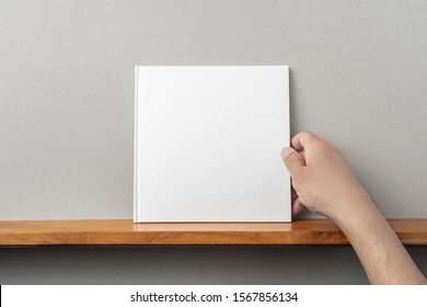 Design concept - man's hand hold square white notebook on bookshelf and grey wall for mockup