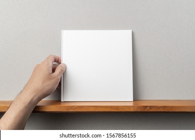 Design concept - man's hand hold square white notebook on bookshelf and grey wall for mockup